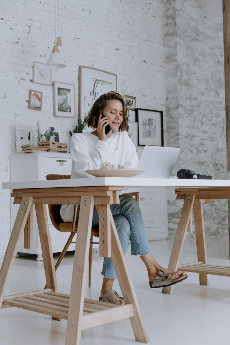 A woman multitasks at home, speaking on the phone while working on her laptop.