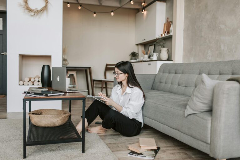 Woman working remotely from a cozy home office, seated on the floor with laptop and notebook.